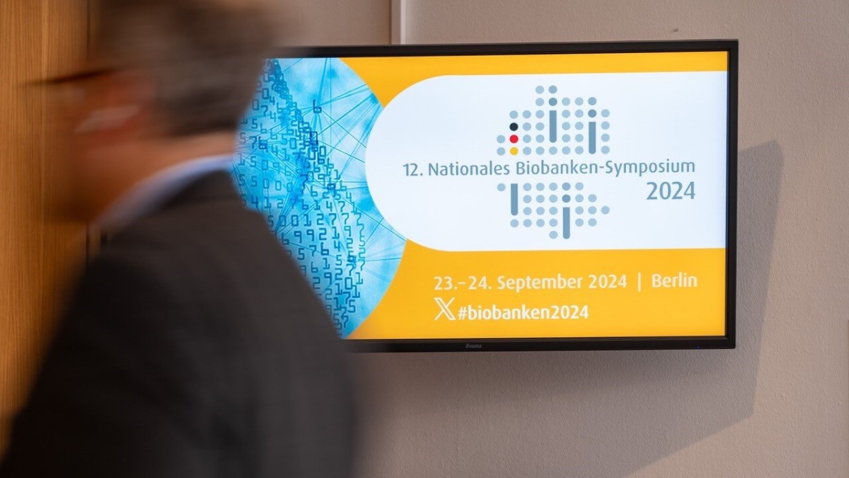 Ein Mann läuft an einem Bildschirm vorbei, auf dem das Banner des 12. Nationalen Biobanken-Symposiums angezeigt wird