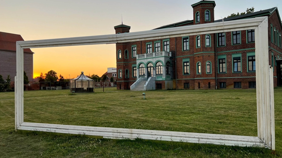 Ein rotes Ziegelgebäude mit türkisen Fensterrahmen. Auf der großen Wiesen davor steht ein leerer weißer Bilderrahmen, der das Gebäude und den Sonnenuntergang daneben einfängt.