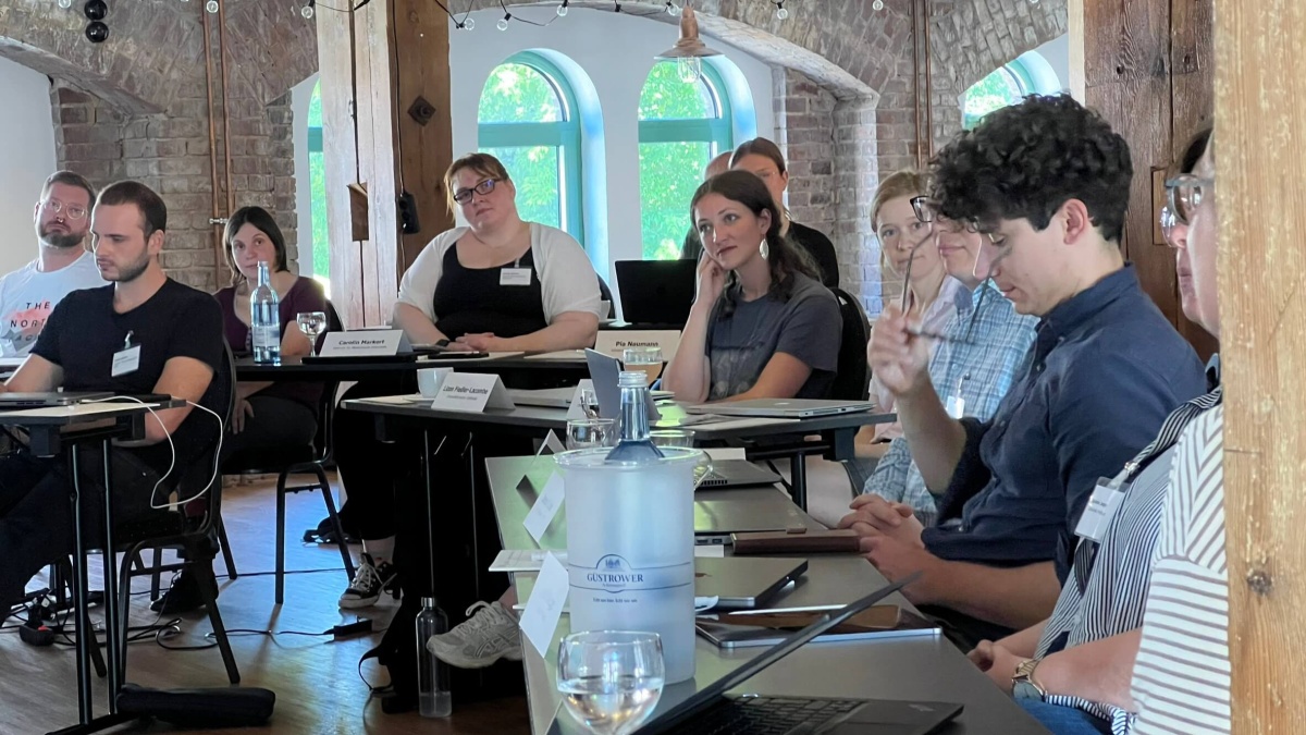 Eine Gruppe Menschen in einem Raum, die an mehreren Tischen sitzen und Computer vor sich stehen haben.