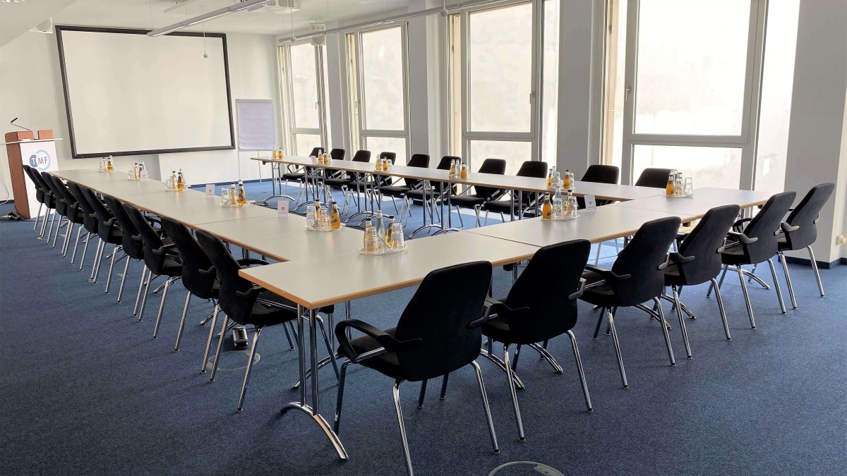 Ein Konferenzraum, in dem mehrere Tische in U-Form platziert sind. Links im Hintergrund sind ein Rednerpult und eine Leinwand zu sehen. Rechts befindet sich eine Fensterfassade.