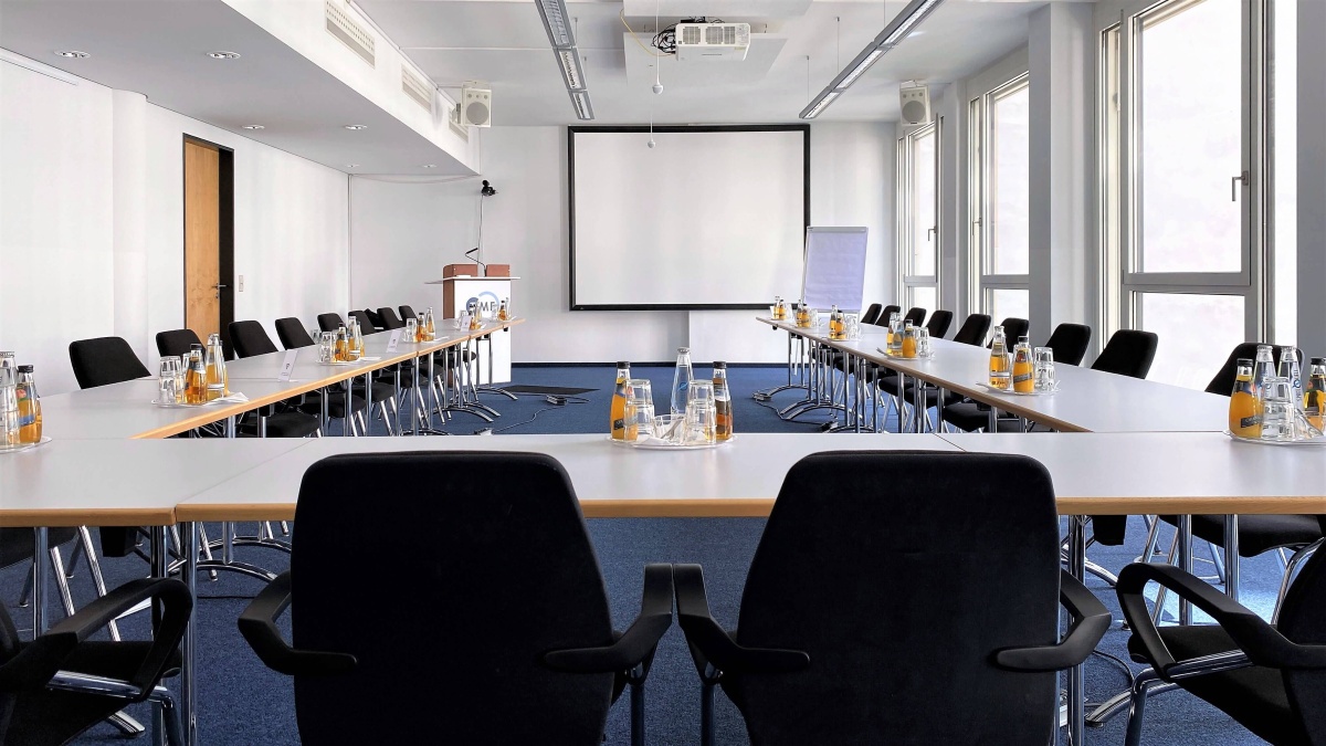 Ein Konferenzraum, in dem mehrere Tische in U-Form platziert sind. Im Hintergrund sind ein Rednerpult und eine Leinwand zu sehen.
