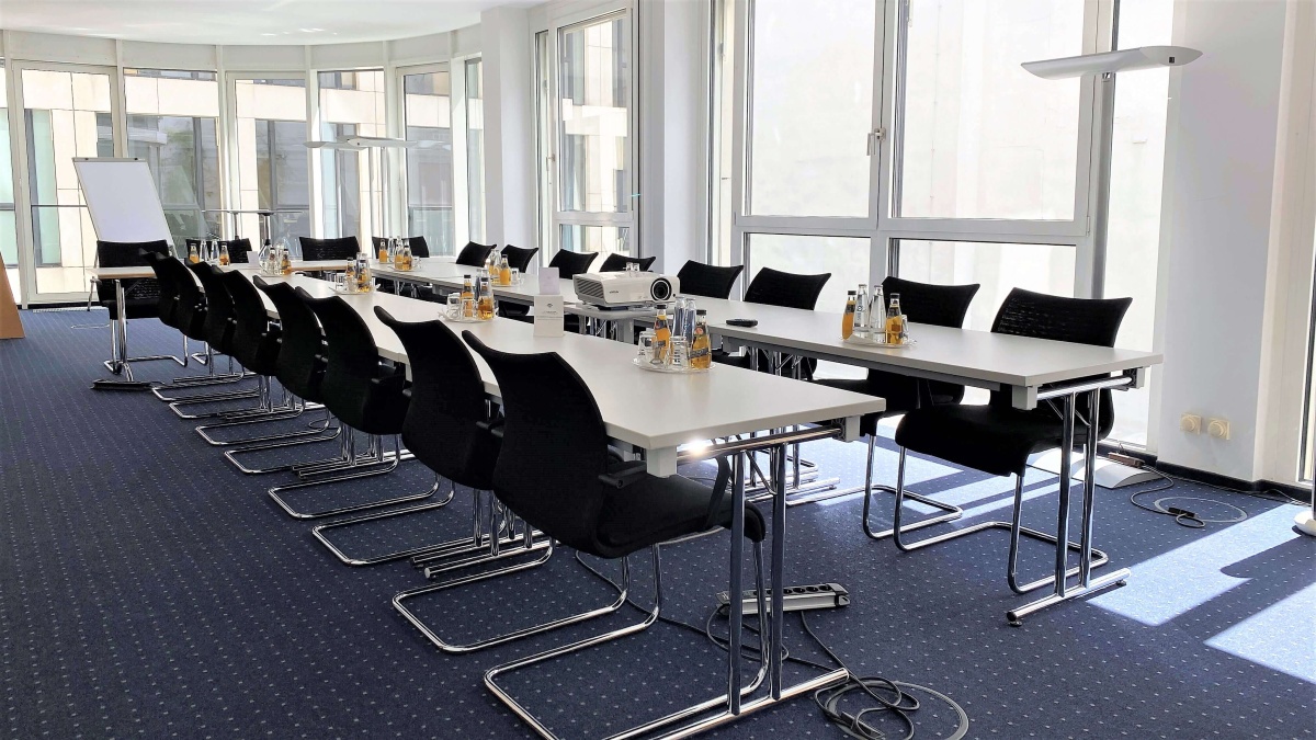 Ein Konferenzraum, in dem mehrere Tische in U-Form platziert sind. In der Mitte steht ein Beamer. Im Hintergrund befindet sich eine lange Fensterfassade.